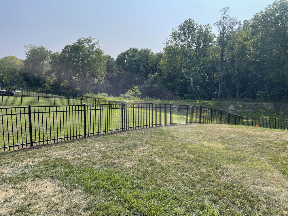 A wide view of black aluminum fences surrounding grassy backyards with trees in the distance.