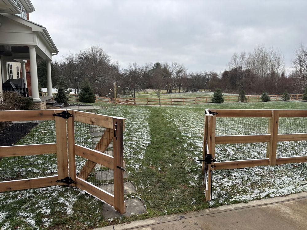 A wooden farm fence with an open gate and wire panels surrounds a grassy yard with a dusting of snow.