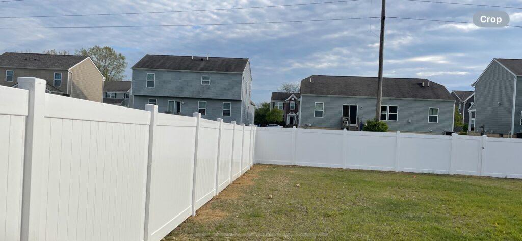 A white vinyl fence with a small gate surrounds a yard in an L-shape. Other houses stand in the background.