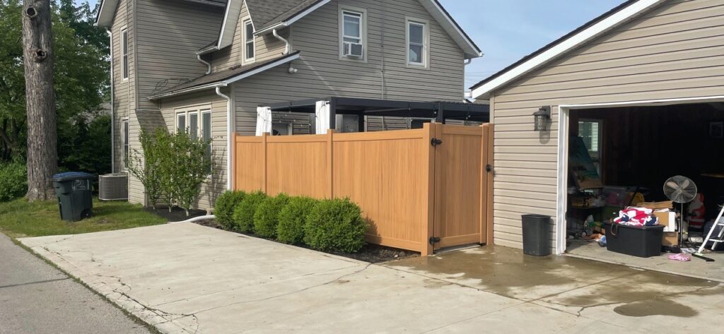 A brown faux wood vinyl fence with a side gate stands between an open garage and a house.