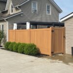 A brown faux wood vinyl fence with a side gate stands between an open garage and a house.
