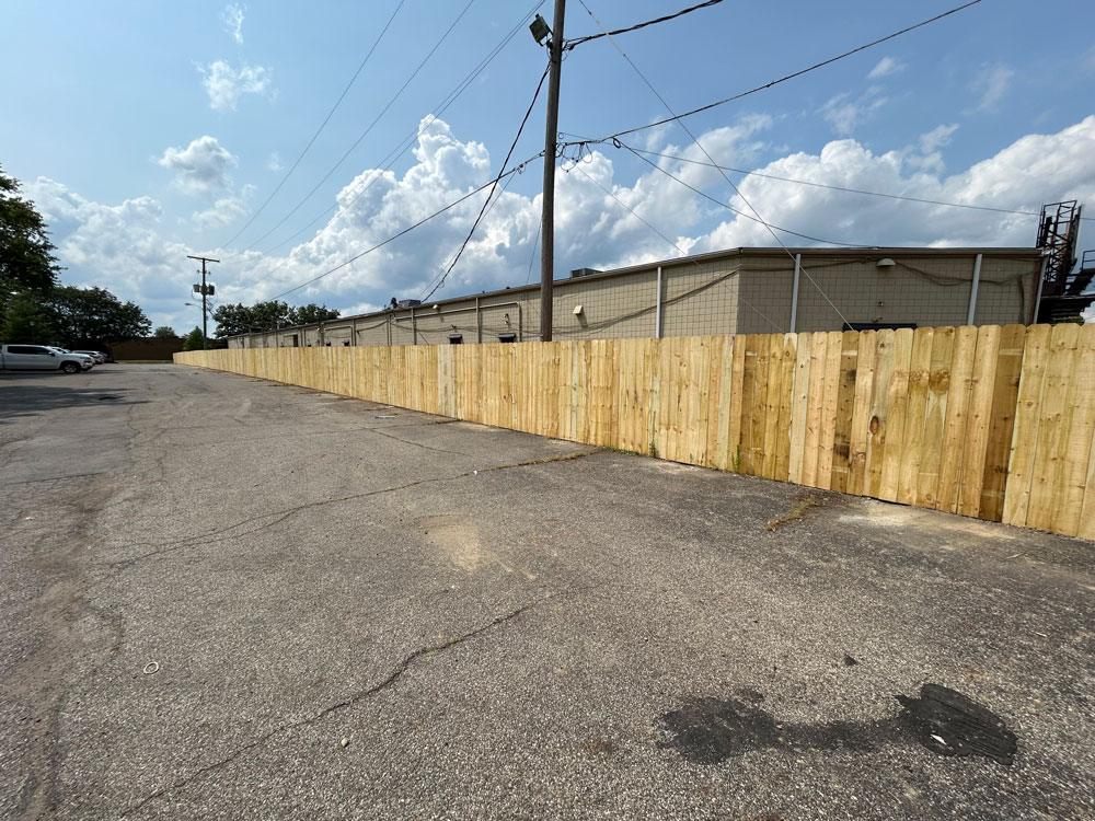 A long commercial wood fence stands between a parking lot and some buildings.